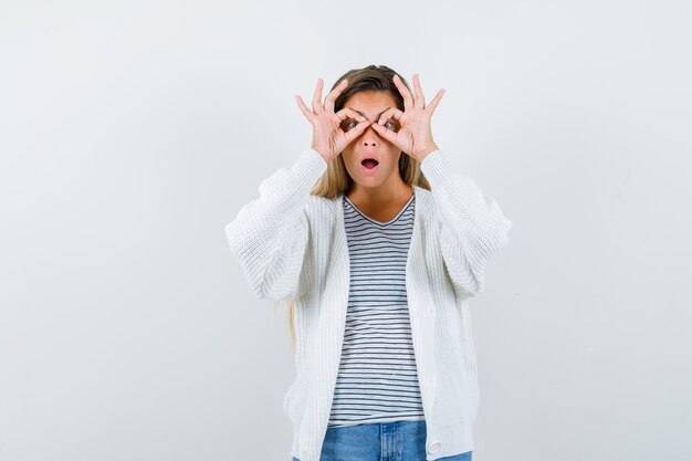 Young woman showing glasses gesture in t-shirt, jacket and looking amazed , front view.