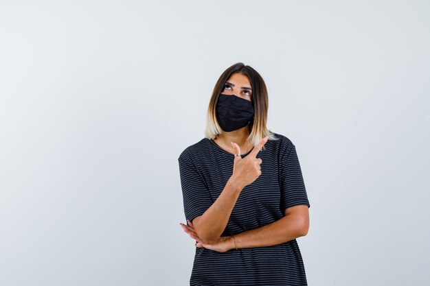 Young woman showing gun gesture, holding hand under elbow in black dress, black mask and looking pensive , front view.