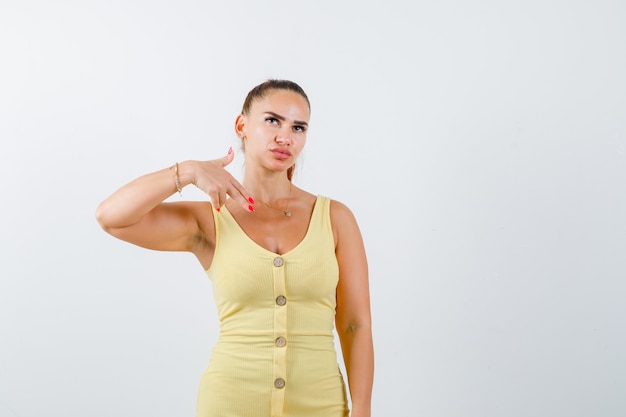 Young woman showing gun gesture, looking up in yellow dress and looking pensive , front view.