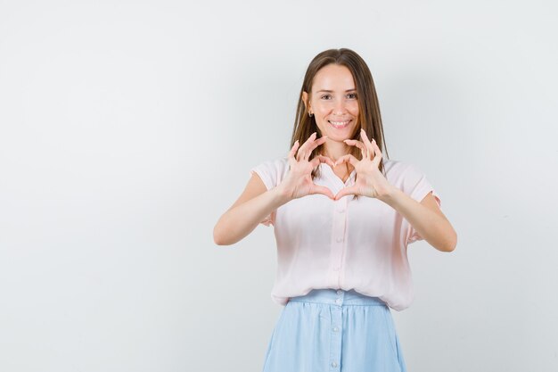 Молодая женщина показывает жест сердца в футболке, юбке и выглядит весело, вид спереди.