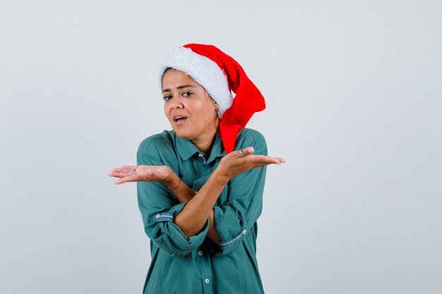 Young woman showing helpless gesture in shirt, Santa hat and looking indecisive , front view.