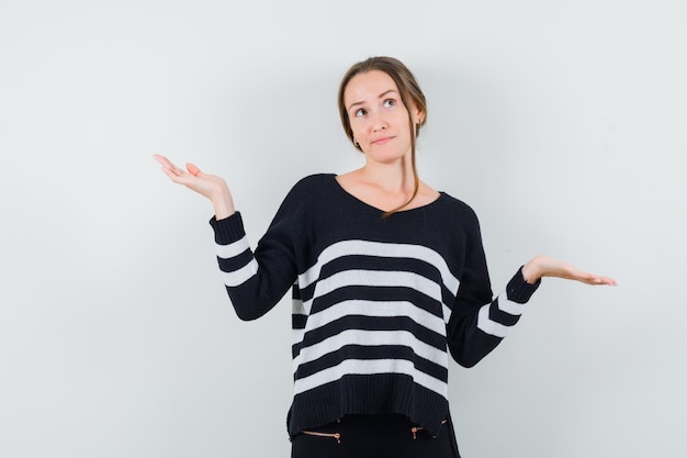 Young woman showing helpless gesture in striped knitwear and black pants and looking pensive