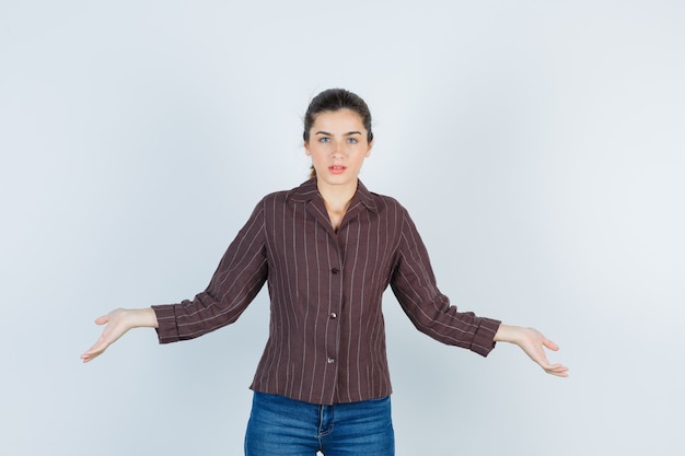 Giovane donna che mostra gesto impotente in camicia a righe, jeans e guardando perplesso, vista frontale.