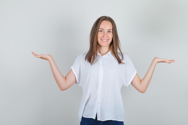 Молодая женщина демонстрирует беспомощный жест в белой футболке, джинсах и выглядит веселой
