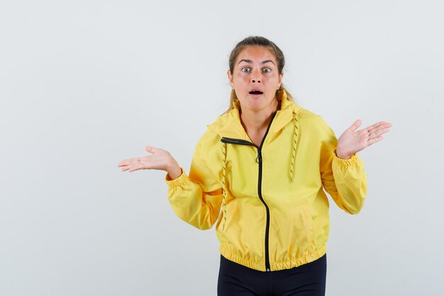 Young woman showing helpless gesture in yellow bomber jacket and black pants and looking baffled