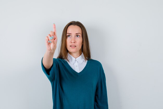 Young woman showing hold on a minute gesture, looking away in sweater over white shirt and looking focused , front view.