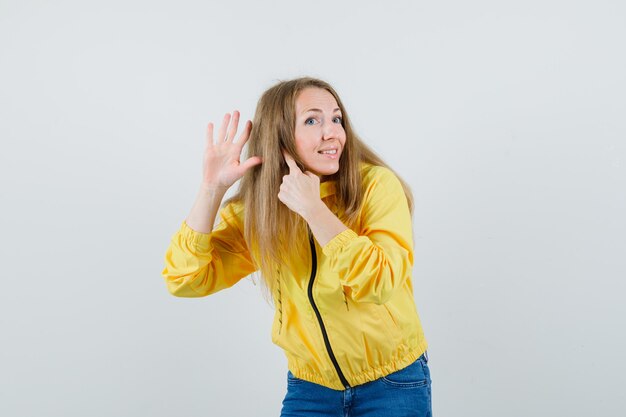 黄色のボンバージャケットとブルージーンズでリスニングジェスチャーを示し、楽観的に見える若い女性。正面図。