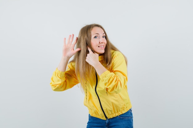 Молодая женщина показывает жест слушания в желтой куртке бомбардировщика и голубых джинсах и выглядит оптимистично. передний план.