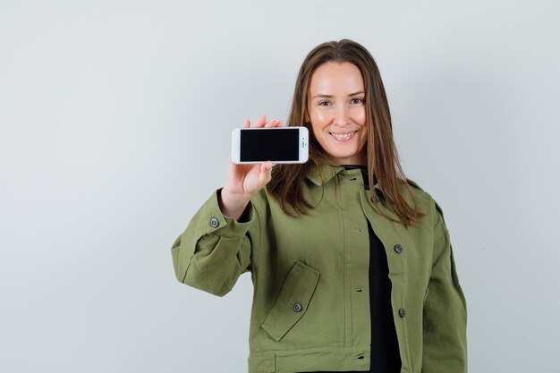 Young woman showing mobile phone in green jacket and looking satisfied , front view.