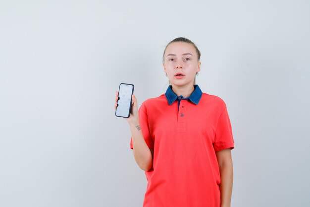 Young woman showing mobile phone in t-shirt and looking puzzled