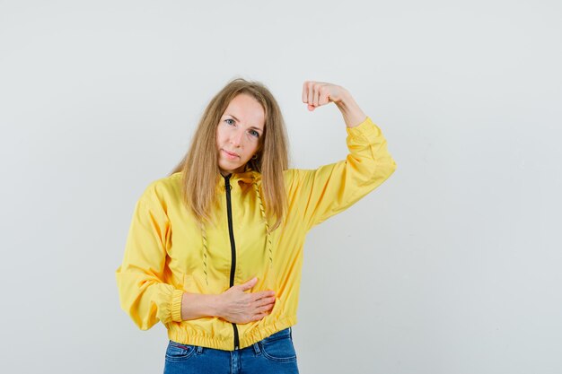 Young woman showing muscles in yellow bomber jacket and blue jean and looking confident, front view.