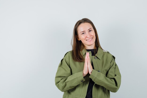 Young woman showing namaste gesture in green jacket and looking glad. front view.