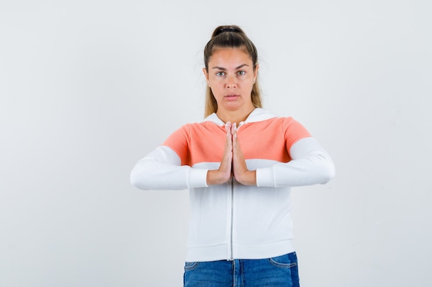 Young woman showing namaste gesture in zip hoodie, jeans and looking calm