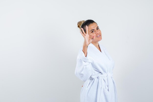 Young woman showing ok gesture in bathrobe and looking blissful