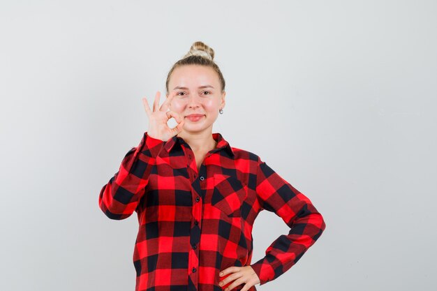 Young woman showing ok gesture in checked shirt and looking cheerful