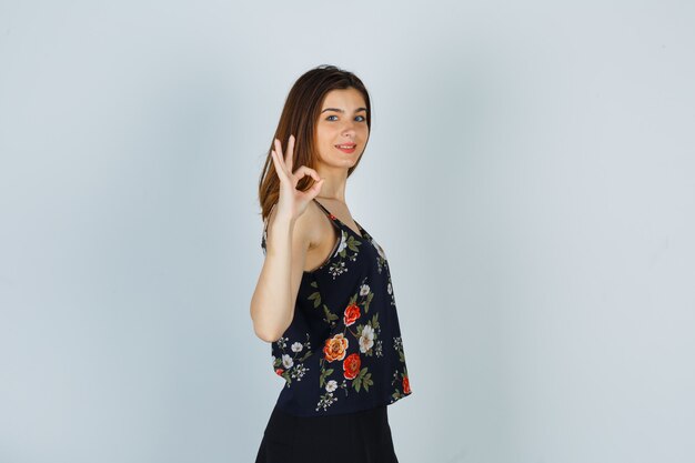 Молодая женщина показывает нормальный жест, боком стоит в цветочном топе и выглядит счастливой.
