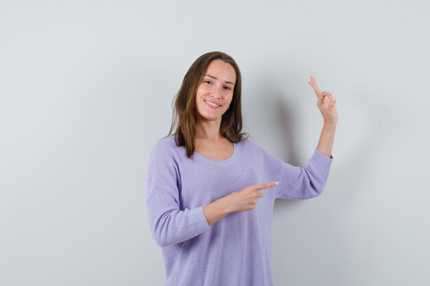 Young woman showing ok gesture while pointing aside in lilac blouse and looking happy 