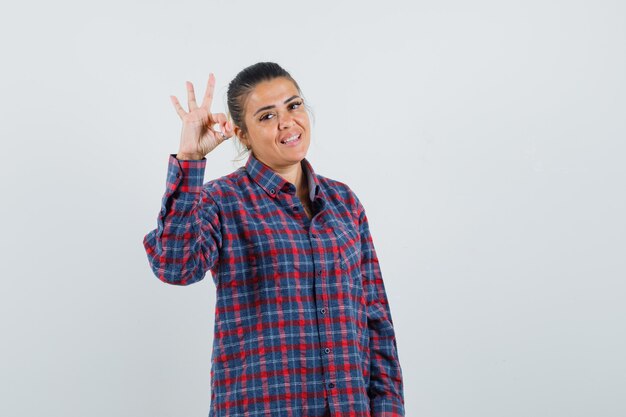 Young woman showing ok sign in checked shirt and looking pretty. front view.