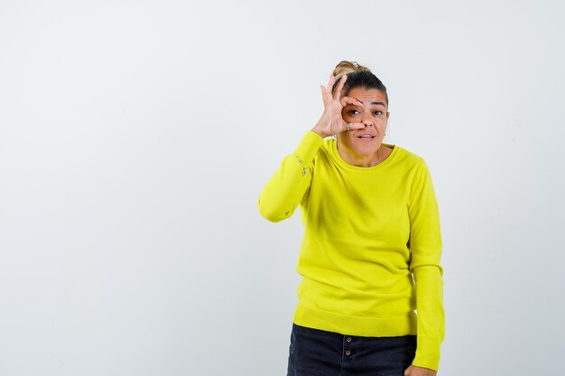 Young woman showing ok sign on eye in yellow sweater and black pants and looking happy 