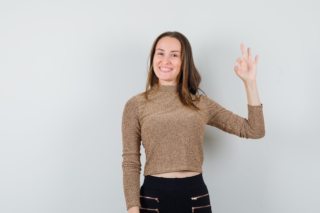 Young woman showing ok sign in gold gilded sweater and black pants and looking happy. front view.