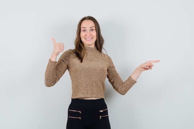 Foto gratuita giovane donna che mostra segno giusto e punta a destra con il dito indice in maglione dorato e pantaloni neri e sembra felice, vista frontale.