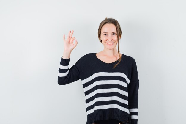 Young woman showing ok sign in striped knitwear and black pants and looking happy