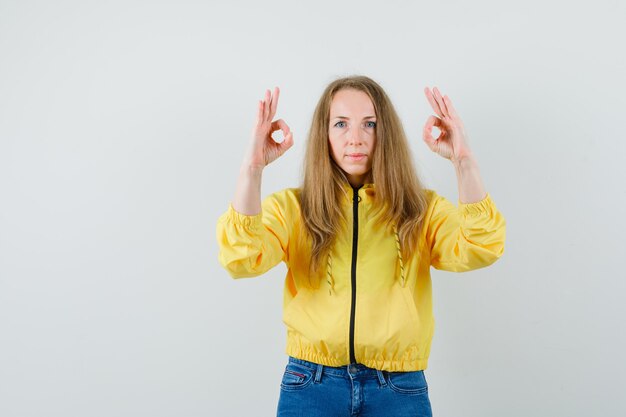 Young woman showing ok sign with both hands in yellow bomber jacket and blue jean and looking serious. front view.