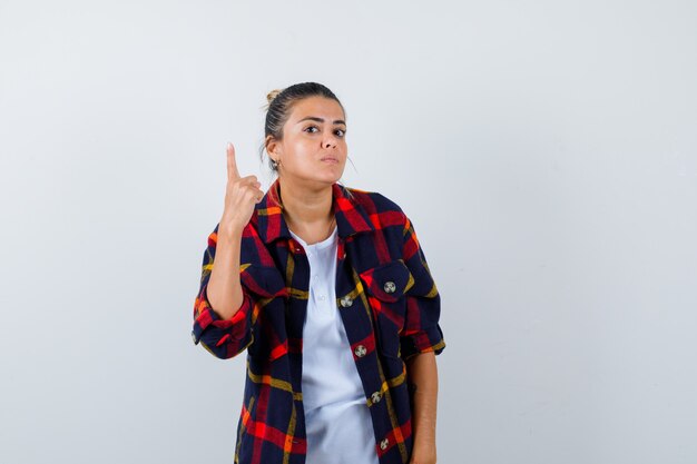 Young woman showing one finger in checkered shirt and looking curious , front view.