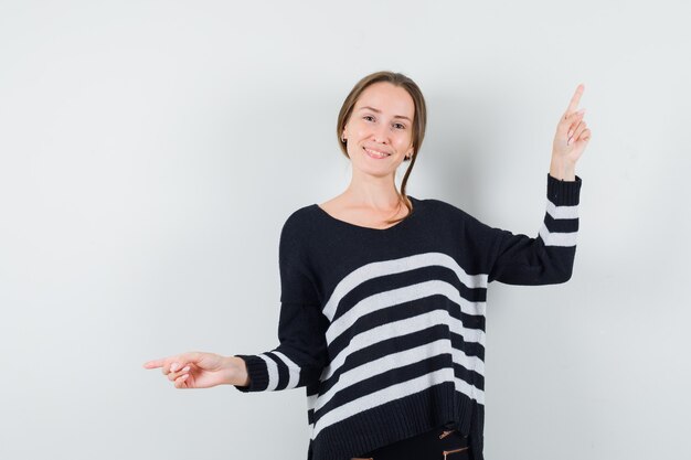 Young woman showing opposite directions with index fingers in striped knitwear and black pants and looking happy