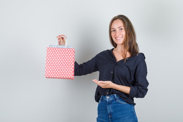 Young woman showing paper bags in black shirt, jeans shorts and looking cheery