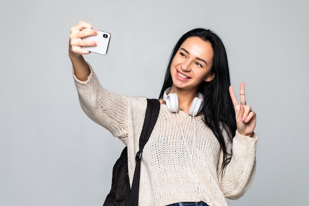 Young woman showing peace gesture while taking selfie on smartphone, isolated on gray wall