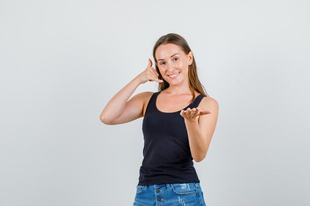 Young woman showing phone gesture in singlet, shorts and looking cheery