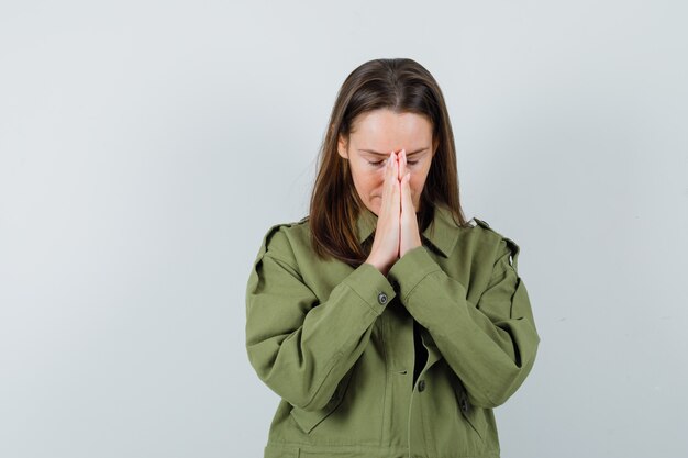 Young woman showing praying gesture in green jacket and looking hopeful. front view.