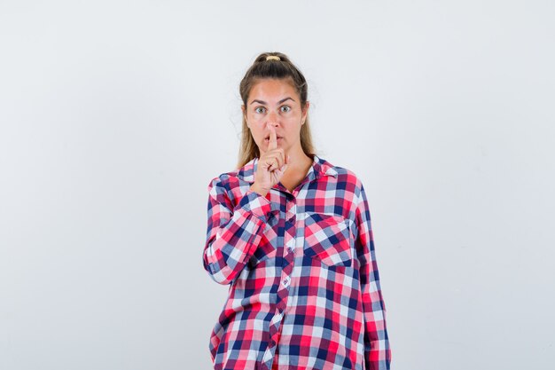 Young woman showing silence gesture in casual shirt and looking careful. front view.