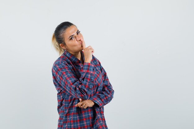Young woman showing silence gesture in checked shirt and looking happy , front view.