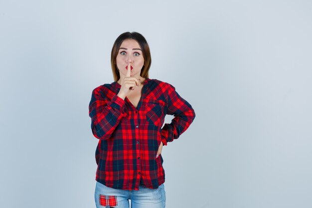 Young woman showing silence gesture in checked shirt and looking scared. front view.
