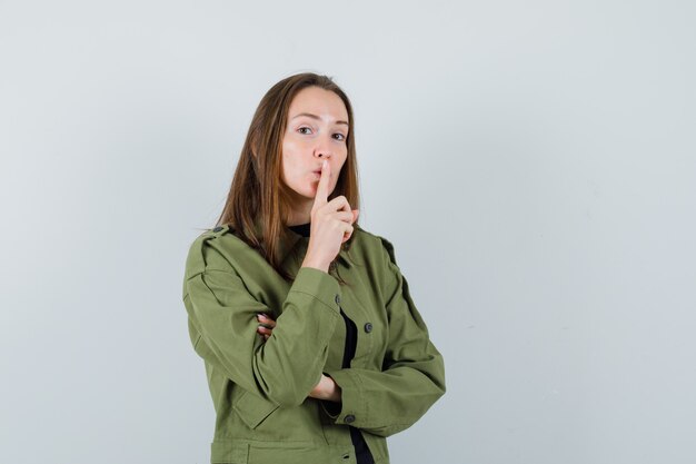 Young woman showing silence gesture in green jacket and looking concentrated. front view.