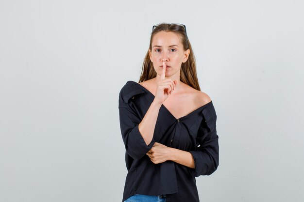 Young woman showing silence gesture in shirt, shorts, glasses and looking careful