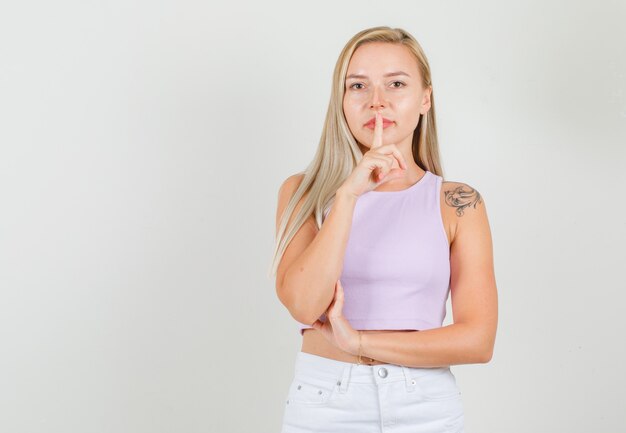 Young woman showing silence gesture in singlet, mini skirt and looking careful.