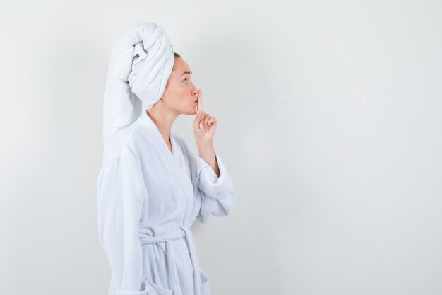 白いバスローブ、タオルで沈黙のジェスチャーを示し、注意深く見ている若い女性。正面図。