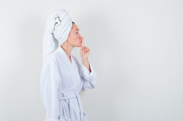 Young woman showing silence gesture in white bathrobe, towel and looking careful , front view.
