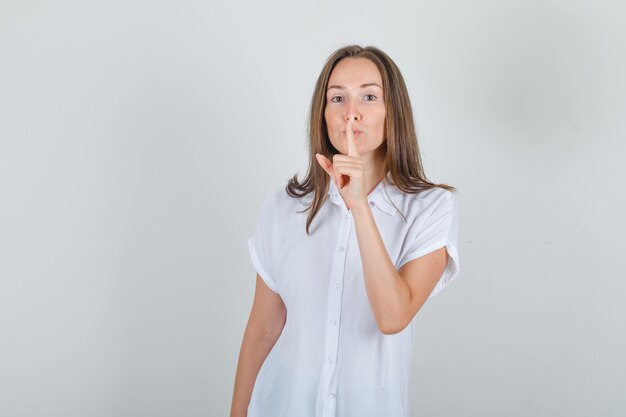 Young woman showing silence gesture in white t-shirt and looking careful