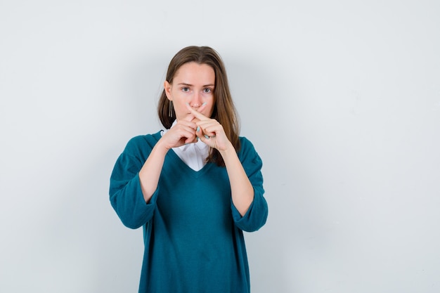 Young woman showing silence gesture with crossed fingers in sweater over white shirt and looking serious. front view.