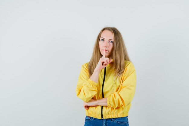 Young woman showing silence gesture in yellow bomber jacket and blue jean and looking serious , front view.