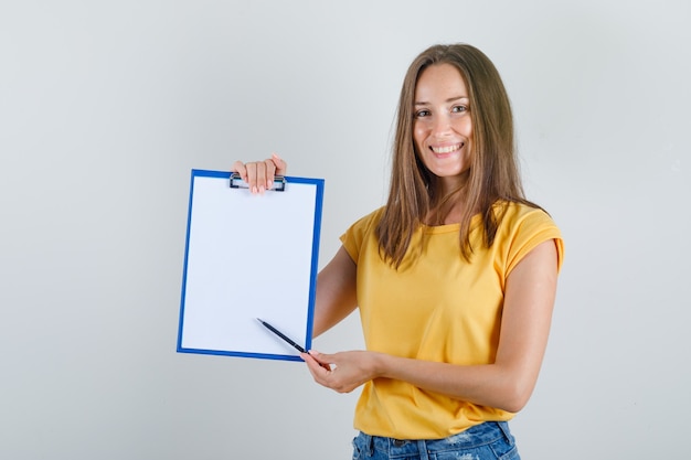 Free photo young woman showing something on clipboard in t-shirt, shorts and looking cheery