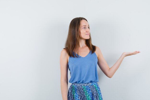 Young woman showing something or welcoming in singlet, skirt and looking confident , front view.