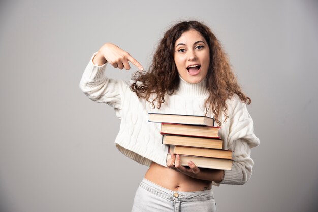 Young woman showing at a stack of books on a gray wall. High quality photo