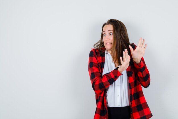 Young woman showing stop gesture in casual clothes and looking scared , front view.