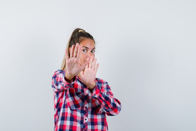 Young woman showing stop gesture in casual shirt and looking scared , front view.