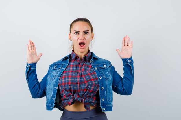 Free photo young woman showing stop gesture in checkered shirt, jean jacket and looking shocked , front view.